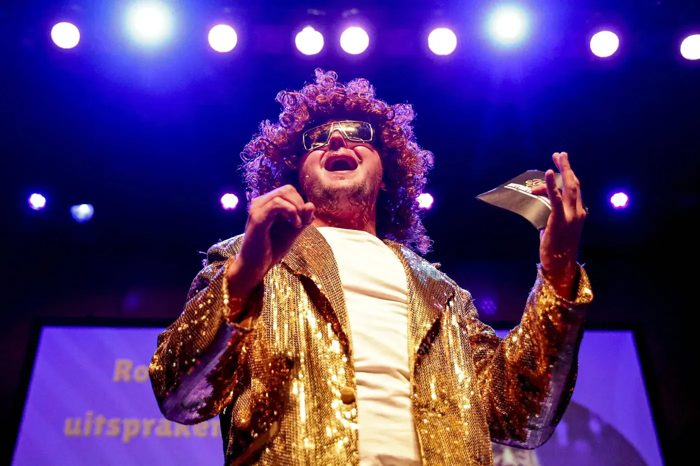 Gast met pruik en glitterjas zingt in de spotlights tijdens de Knallende Karaoke